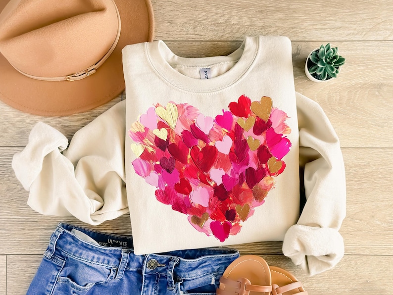 May include: A cream-colored sweatshirt with a large heart design made of painted pink, red, and gold hearts. The sweatshirt is laid out on a wooden surface with a brown hat, blue jeans, and sandals.