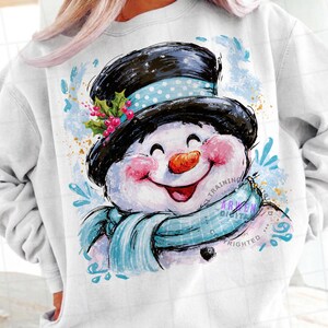 May include: A white sweatshirt featuring a graphic of a smiling snowman wearing a black top hat with a light blue polka dot band, a light blue scarf, and holly berries. The design includes watercolor-style blue splashes and text.
