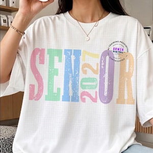 Puede incluir: Una camiseta blanca con un patr&oacute;n de cuadr&iacute;cula de fondo y la palabra SENIOR 2027 impresa en letras grandes, desgastadas y multicolores. Las letras son de color rosa, verde, azul, amarillo y morado. Se observa un peque&ntilde;o logotipo circular.