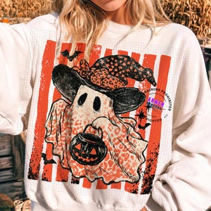 May include: A white crewneck sweatshirt featuring a graphic of a ghost wearing a black star-patterned witch hat and holding a jack-o'-lantern bucket. The ghost has an orange leopard print pattern and is set against a background of vertical orange stripes.