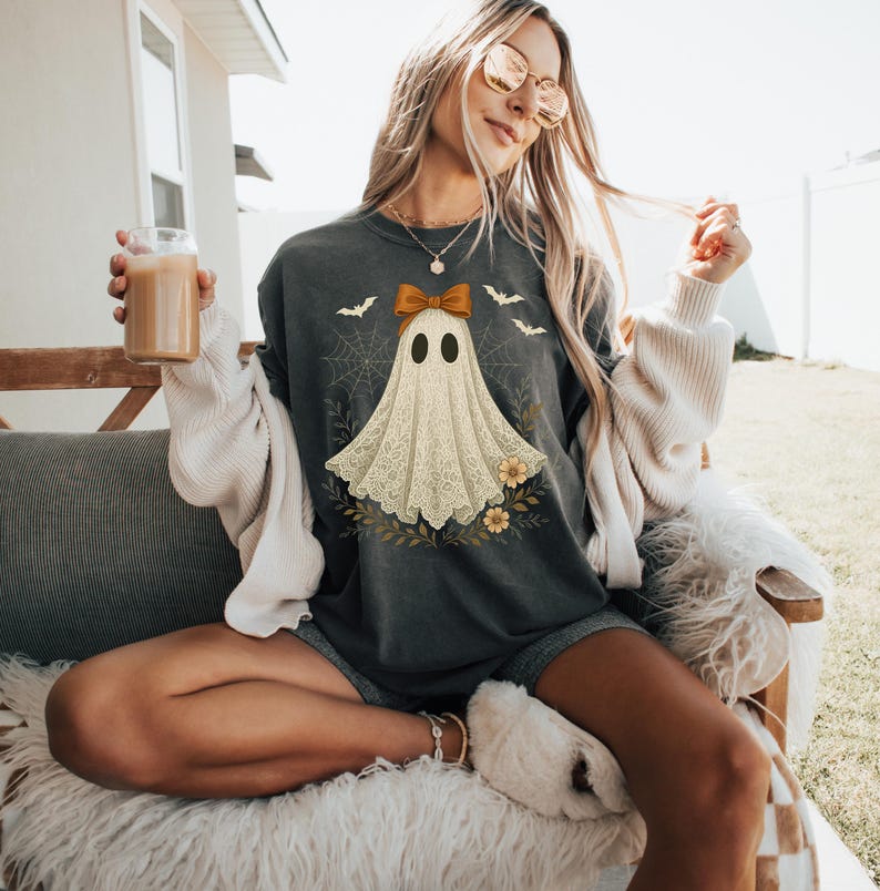 May include: Dark gray sweatshirt featuring a ghost design with a bow, bats, and flowers. The ghost has a lace-like appearance. The design is centered on the front of the shirt, perfect for Halloween or spooky season.