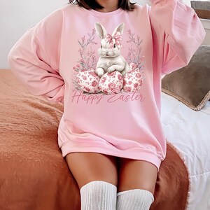 May include: A pink sweatshirt with a white bunny rabbit wearing a pink bow sitting on a nest of pink and white floral patterned Easter eggs. The text "Happy Easter" is printed below the bunny.