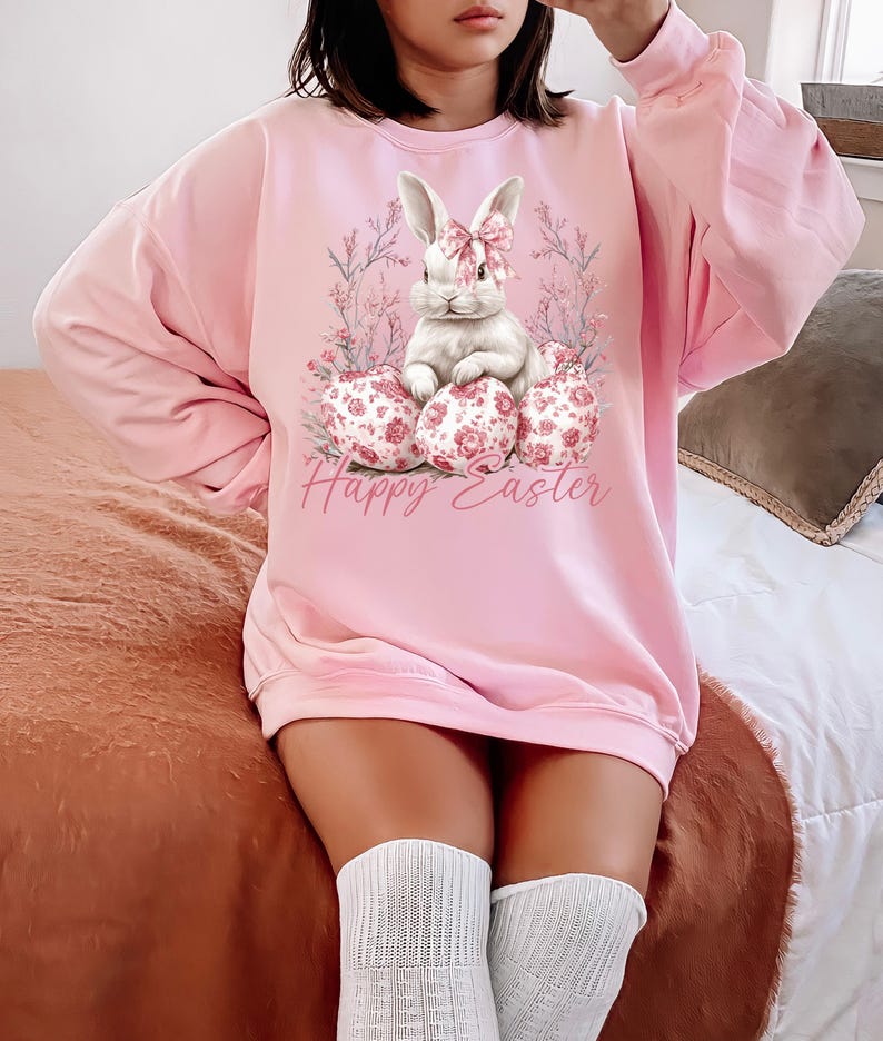 May include: A pink sweatshirt with a white bunny rabbit wearing a pink bow sitting on a nest of pink and white floral patterned Easter eggs. The text "Happy Easter" is printed below the bunny.