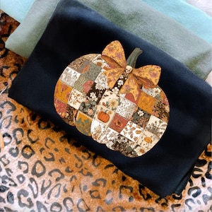 May include: A black sweatshirt featuring a patchwork pumpkin design with a bow and skull detail. The pumpkin is composed of various floral and patterned squares in autumnal colors. The sweatshirt is layered with other folded fabrics and a leopard print.