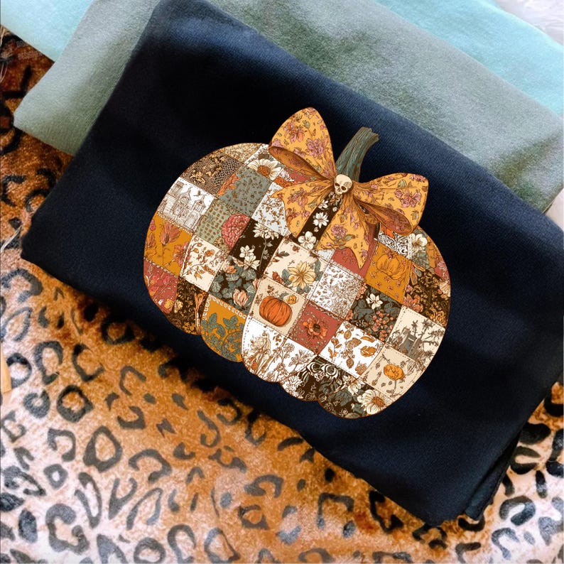 May include: A black sweatshirt featuring a patchwork pumpkin design with a bow and skull detail. The pumpkin is composed of various floral and patterned squares in autumnal colors. The sweatshirt is layered with other folded fabrics and a leopard print.