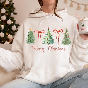 May include: White hoodie with a watercolor design of Christmas trees and red bows, with the words "Merry Christmas" in red script. A festive holiday sweatshirt for the Christmas season.