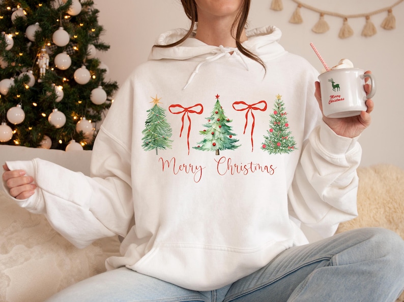 May include: White hoodie with a watercolor design of Christmas trees and red bows, with the words "Merry Christmas" in red script. A festive holiday sweatshirt for the Christmas season.