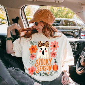 K&ouml;nnte beinhalten: Cremefarbenes T-Shirt mit einem Halloween-Motiv. Das Design zeigt einen Cartoon-Geist mit einer Leopardenmuster-Schleife, umgeben von orangefarbenen und rosa Blumen, und den Worten "Spooky Season."