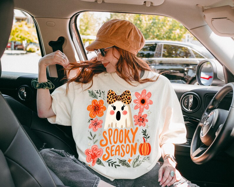 K&ouml;nnte beinhalten: Cremefarbenes T-Shirt mit einem Halloween-Motiv. Das Design zeigt einen Cartoon-Geist mit einer Leopardenmuster-Schleife, umgeben von orangefarbenen und rosa Blumen, und den Worten "Spooky Season."