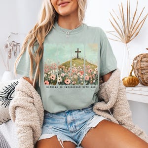 May include: A light blue t-shirt with a floral design and a cross on a hill. The text "Nothing is impossible with God" is printed on the shirt.