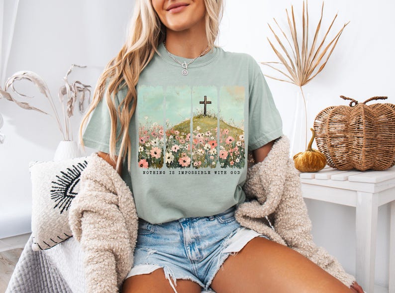 May include: A light blue t-shirt with a floral design and a cross on a hill. The text "Nothing is impossible with God" is printed on the shirt.