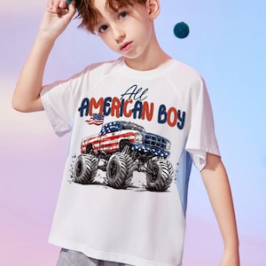 May include: White t-shirt with a graphic of a monster truck decorated with the American flag and the text "All AMERICAN BOY". The shirt has blue accents on the sides and is worn by a person wearing a blue baseball cap.