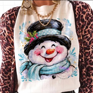 May include: A white t-shirt featuring a graphic illustration of a smiling snowman wearing a black top hat with a light blue polka dot band, a light blue scarf, and a sprig of holly with red berries, layered under a brown and black leopard print cardigan.