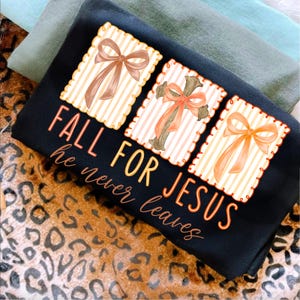 May include: Black sweatshirt with the text "FALL FOR JESUS he never leaves." The design includes three rectangular images with bows and a cross. The bows are in shades of brown and orange. The background is a leopard print.