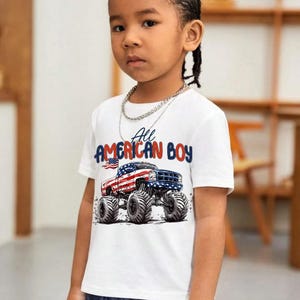 May include: A white t-shirt with the text "All AMERICAN BOY" and a graphic of a monster truck decorated with the American flag. The shirt is worn by a young person with dark hair and a silver chain necklace.