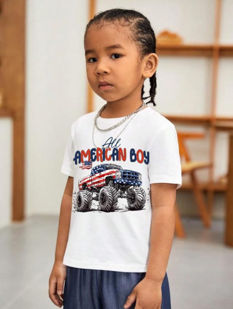 May include: A white t-shirt with the text "All AMERICAN BOY" and a graphic of a monster truck decorated with the American flag. The shirt is worn by a young person with dark hair and a silver chain necklace.