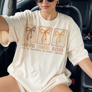 May include: Cream-colored t-shirt featuring three square designs with bows in shades of brown and orange. The text "Grateful Thankful Blessed" is printed below the bows, with the phrase "God is faithful in every season."