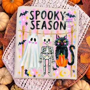 K&ouml;nnte beinhalten: Ein rechteckiger Druck mit dem Text SPOOKY SEASON in schwarzen Buchstaben. Darunter h&auml;ngen ein wei&szlig;es Gespenst, ein Skelett und eine schwarze Katze mit einem orangefarbenen K&uuml;rbis an einer W&auml;scheleine mit Holzklammern vor einem rosa gestreiften Hintergrund.