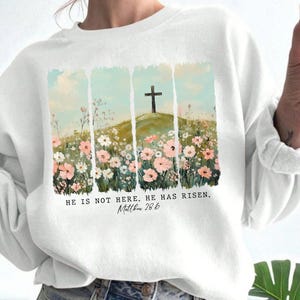 May include: White sweatshirt with a floral design and a cross on a hill. The text "He is not here. He has risen. Matthew 28:6" is printed below the design.