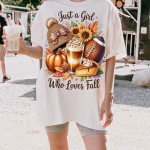K&ouml;nnte beinhalten: Cremefarbenes T-Shirt mit einem herbstlichen Motiv. Das Design umfasst einen K&uuml;rbis, Sonnenblumen, einen Football, eine M&uuml;tze, ein Kaffeegetr&auml;nk und ein St&uuml;ck Kuchen. Der Text lautet "Just a Girl Who Loves Fall."