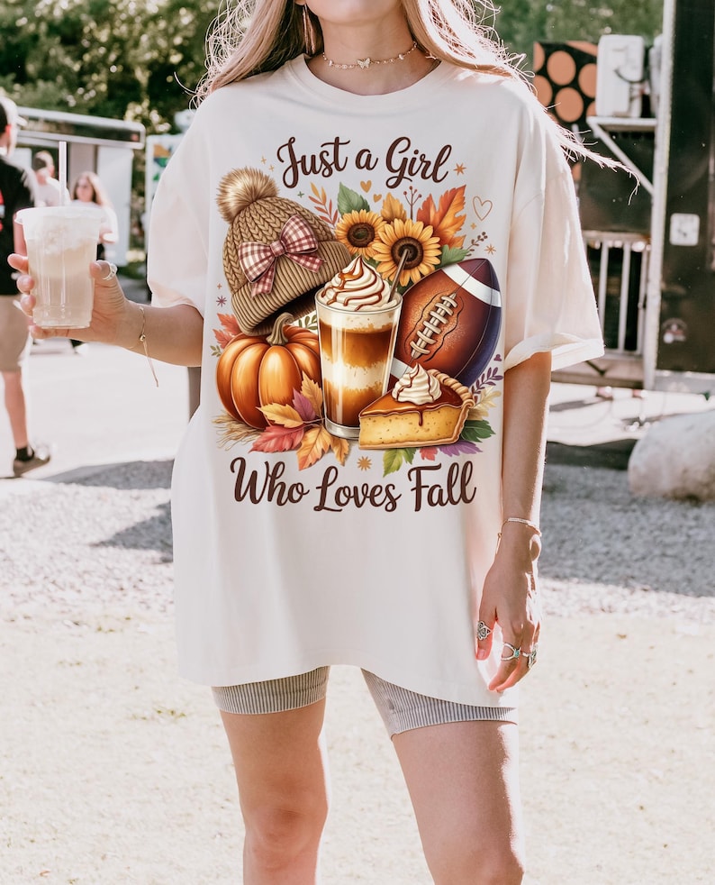 K&ouml;nnte beinhalten: Cremefarbenes T-Shirt mit einem herbstlichen Motiv. Das Design umfasst einen K&uuml;rbis, Sonnenblumen, einen Football, eine M&uuml;tze, ein Kaffeegetr&auml;nk und ein St&uuml;ck Kuchen. Der Text lautet "Just a Girl Who Loves Fall."