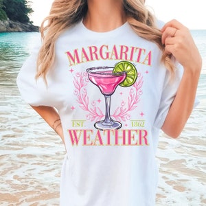 May include: White t-shirt featuring a pink margarita glass graphic and the text "Margarita Weather" with "Est. 1862" below the graphic.