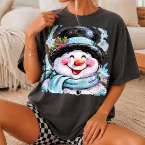 May include: A charcoal gray short-sleeve t-shirt featuring a graphic print of a smiling snowman wearing a black top hat with a light blue polka dot band and a matching light blue scarf. The snowman has rosy cheeks, a carrot nose, and holly berries.