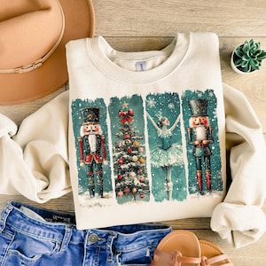 May include: Cream-colored sweatshirt featuring a festive design with nutcrackers, a Christmas tree, and a ballerina. The graphic is set against a teal background with falling snow. The sweatshirt is paired with blue jeans, sandals, and a tan hat.