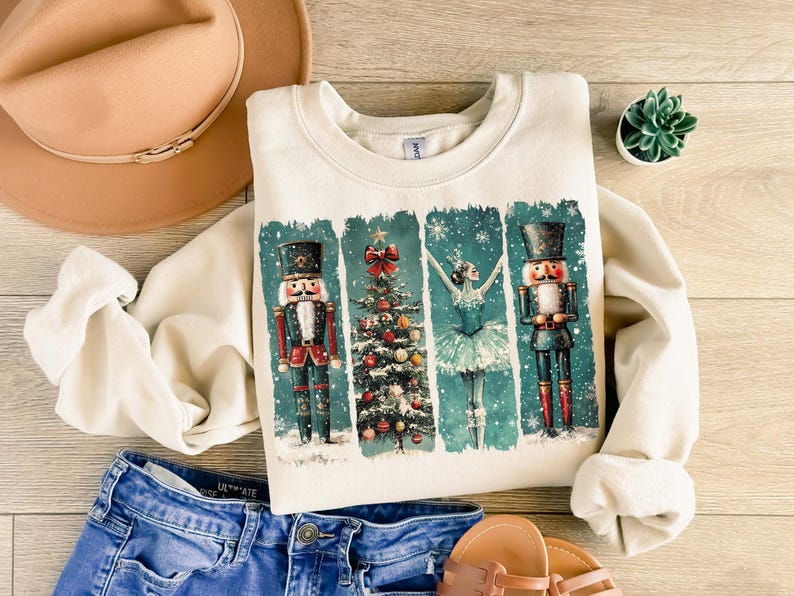 May include: Cream-colored sweatshirt featuring a festive design with nutcrackers, a Christmas tree, and a ballerina. The graphic is set against a teal background with falling snow. The sweatshirt is paired with blue jeans, sandals, and a tan hat.
