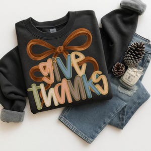May include: A black sweatshirt with the words "GIVE THANKS" in colorful letters and a brown bow design. The sweatshirt is paired with blue jeans, a gray beanie, pine cones, and a candle.
