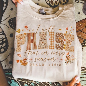 May include: A cream-coloured sweatshirt featuring the phrase "I will PRAISE Him in every season". The word "PRAISE" is designed with floral and plaid patterns. The text "PSALM 146:2" is printed at the bottom of the garment.