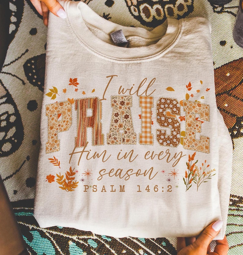 May include: A cream-coloured sweatshirt featuring the phrase "I will PRAISE Him in every season". The word "PRAISE" is designed with floral and plaid patterns. The text "PSALM 146:2" is printed at the bottom of the garment.