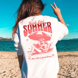 May include: White sweatshirt with a pink graphic design of a woman reading a book under an umbrella on a beach. The text on the shirt says "Book Girl Summer" and "Dive into a good book."