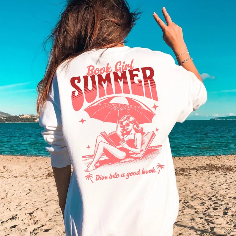 May include: White sweatshirt with a pink graphic design of a woman reading a book under an umbrella on a beach. The text on the shirt says "Book Girl Summer" and "Dive into a good book."