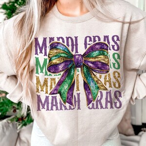 Puede incluir: Una sudadera beige con el texto "Mardi Gras" repetido cuatro veces en un dise&ntilde;o en capas. Un gran lazo de purpurina morado, verde y dorado est&aacute; centrado en la sudadera.