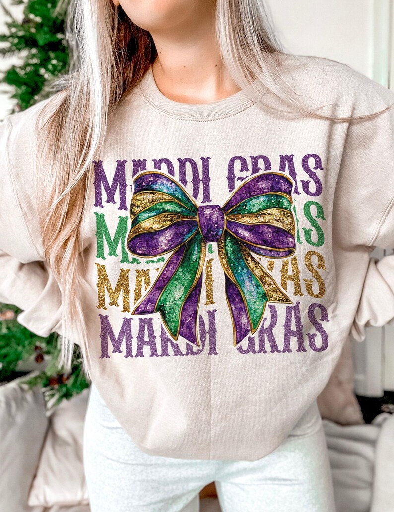 Puede incluir: Una sudadera beige con el texto "Mardi Gras" repetido cuatro veces en un dise&ntilde;o en capas. Un gran lazo de purpurina morado, verde y dorado est&aacute; centrado en la sudadera.