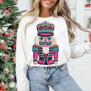 May include: White long-sleeve sweatshirt featuring a colorful nutcracker design. The nutcracker has a crown, a blue mustache, and a pink and teal uniform. The sweatshirt is a festive holiday item.