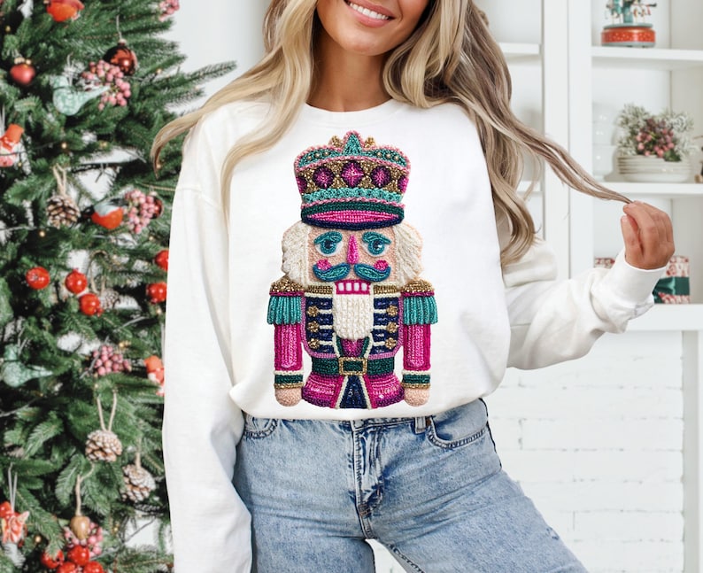 May include: White long-sleeve sweatshirt featuring a colorful nutcracker design. The nutcracker has a crown, a blue mustache, and a pink and teal uniform. The sweatshirt is a festive holiday item.