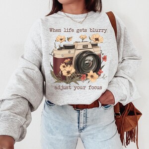 May include: Gray sweatshirt with a vintage camera graphic and the text "When life gets blurry adjust your focus". The camera is surrounded by flowers in shades of yellow, red, and orange.