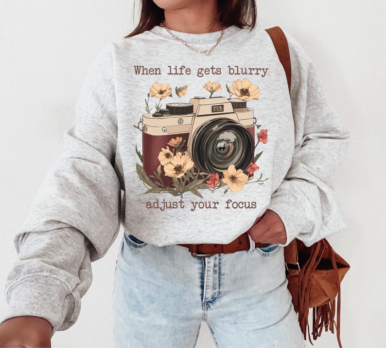 May include: Gray sweatshirt with a vintage camera graphic and the text "When life gets blurry adjust your focus". The camera is surrounded by flowers in shades of yellow, red, and orange.