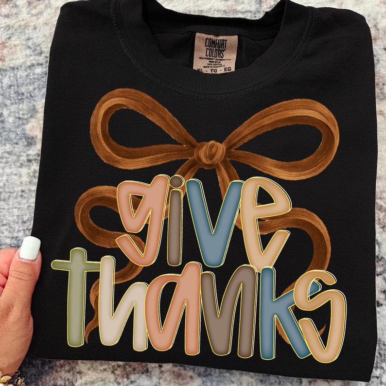 May include: Black t-shirt with a brown bow design and the words "GIVE THANKS" in colorful letters. The letters have a gold outline. The shirt is a Comfort Colors brand, size XL-TG-EG.