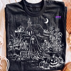 May include: A black t-shirt featuring a white graphic print of a spooky scene with a church, a crescent moon, corn stalks, pumpkins, and a ghost. A rectangular sign in the foreground displays the text "LIVE HERE NOW" in a bold, white font.