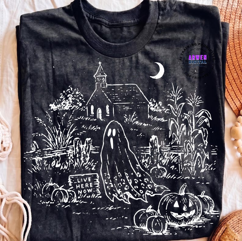 May include: A black t-shirt featuring a white graphic print of a spooky scene with a church, a crescent moon, corn stalks, pumpkins, and a ghost. A rectangular sign in the foreground displays the text "LIVE HERE NOW" in a bold, white font.
