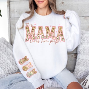 以下が含まれることがあります： ピンクとゴールドのグリッター文字で「This Mama loves her peeps」と書かれた白いスウェットシャツ。袖にはゴールドのグリッターのウサギのデザインが施されています。
