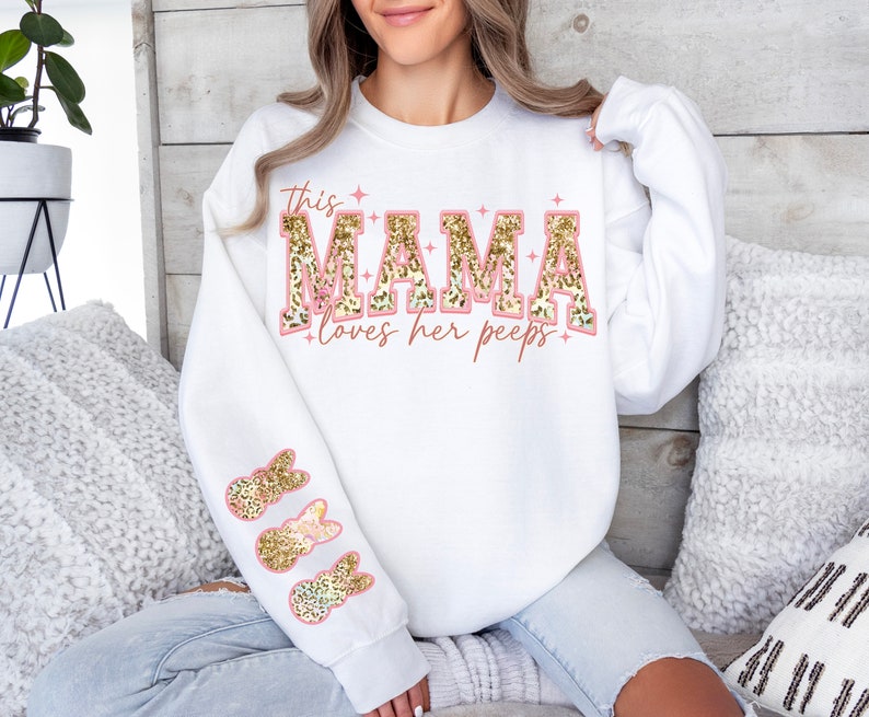 以下が含まれることがあります： ピンクとゴールドのグリッター文字で「This Mama loves her peeps」と書かれた白いスウェットシャツ。袖にはゴールドのグリッターのウサギのデザインが施されています。