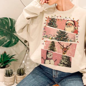 May include: Cream-colored sweatshirt featuring a festive Christmas design. The graphic includes Santa Claus, a reindeer, a snowman, and Christmas trees, all set against a pink backdrop. The design is framed with string lights.