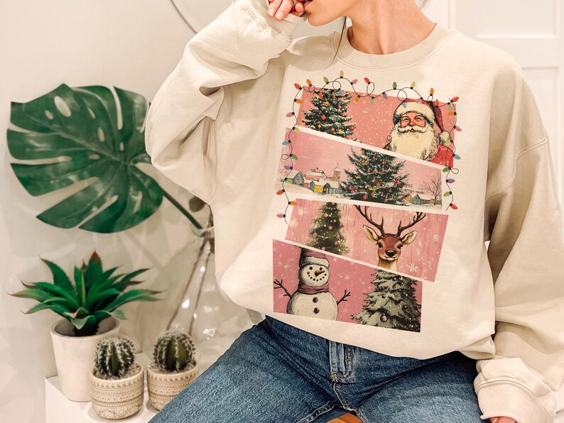 May include: Cream-colored sweatshirt featuring a festive Christmas design. The graphic includes Santa Claus, a reindeer, a snowman, and Christmas trees, all set against a pink backdrop. The design is framed with string lights.
