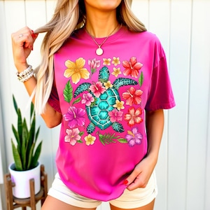 May include: A pink t-shirt featuring a graphic of a sea turtle surrounded by colourful flowers. The turtle is turquoise and green, and the flowers are pink, yellow and orange.