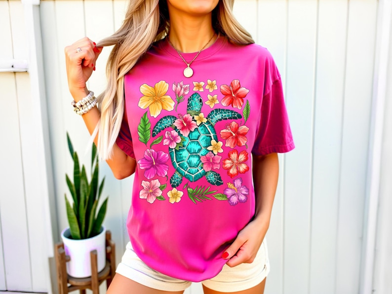 May include: A pink t-shirt featuring a graphic of a sea turtle surrounded by colourful flowers. The turtle is turquoise and green, and the flowers are pink, yellow and orange.