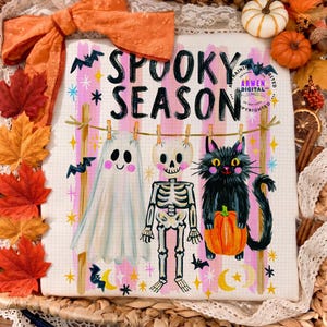 K&ouml;nnte beinhalten: Ein wei&szlig;er Stoffdruck mit dem schwarzen Schriftzug SPOOKY SEASON, der ein Gespenst, ein Skelett und eine schwarze Katze mit einem K&uuml;rbis an einer W&auml;scheleine zeigt. Der Hintergrund ist mit rosa Streifen, gelben Sternen und Flederm&auml;usen verziert.